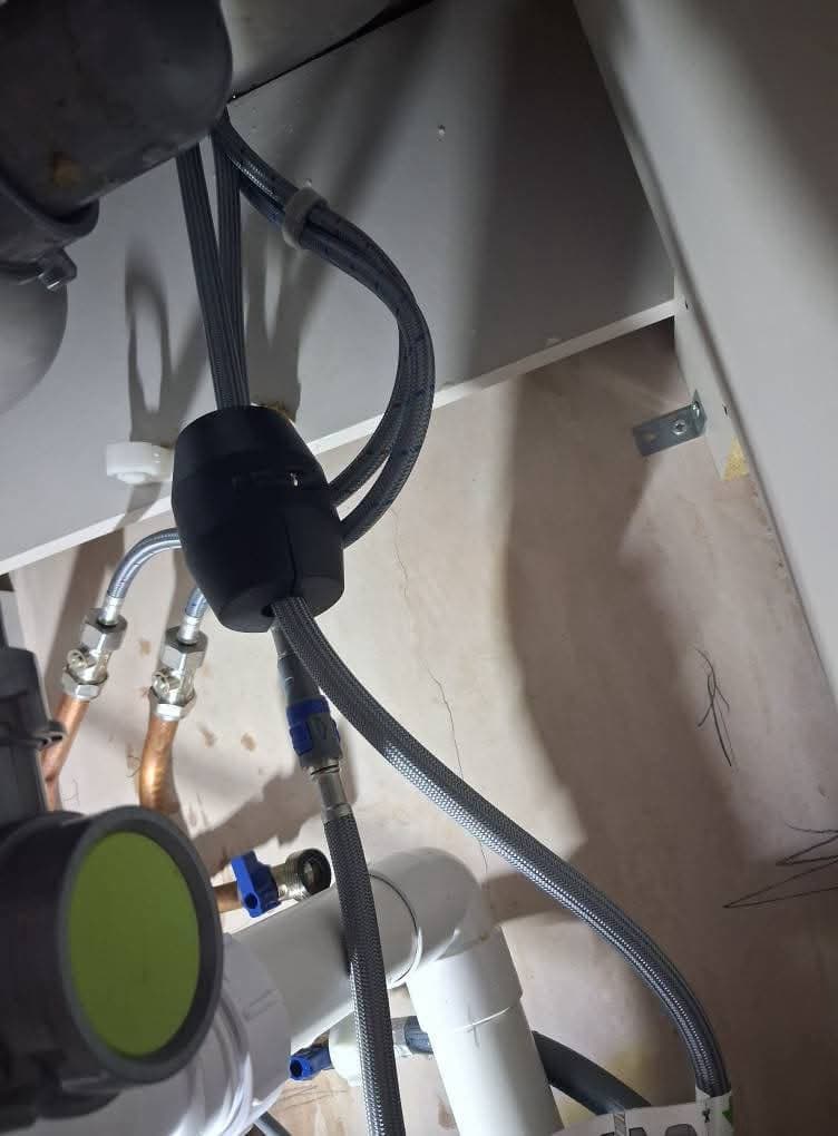 Under a sink: grey appliance hoses, a black anti-siphon fitting and waste pipework